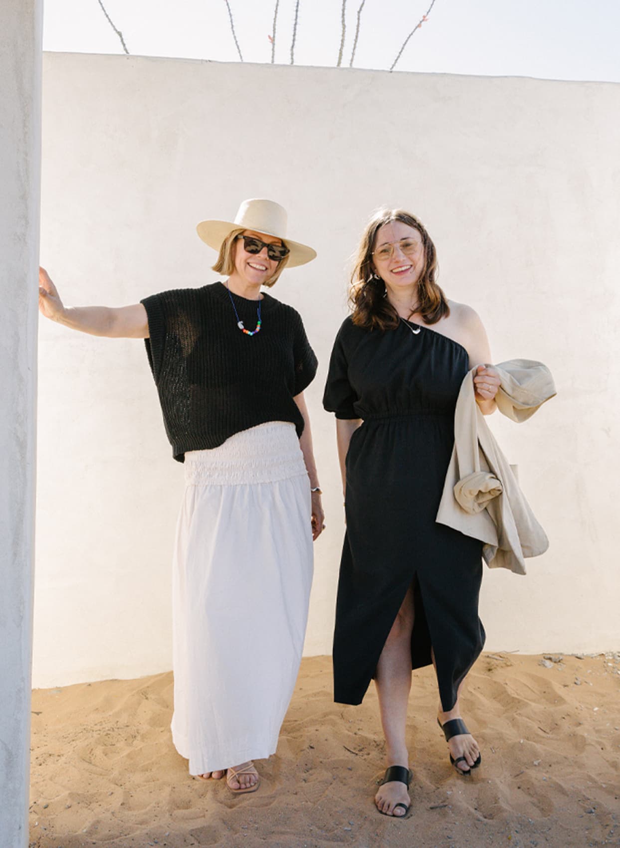 Two woman, one wearing black and white and the other wearing all black, stand on sand outdoors smiling