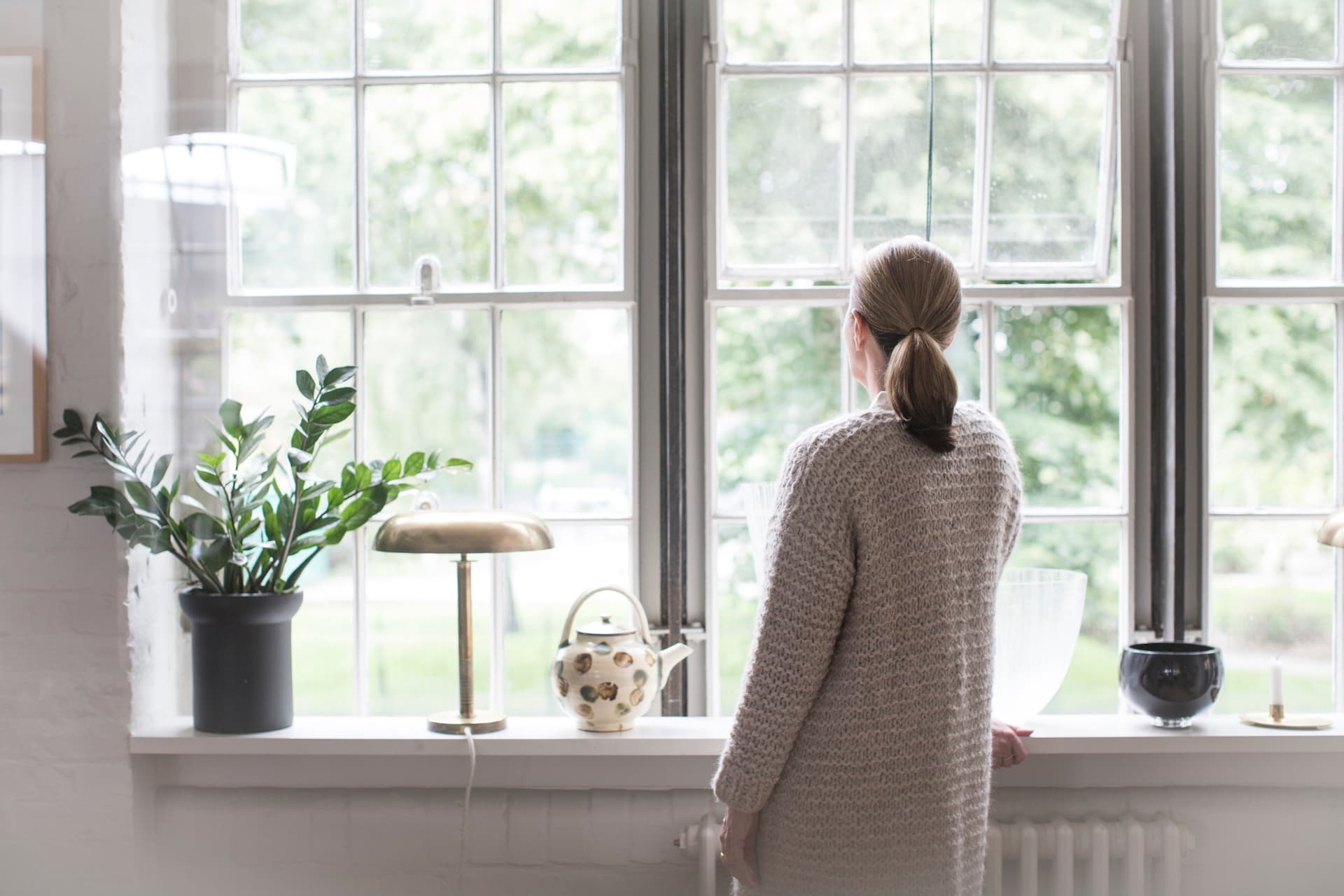 Ilse Crawford stands facing away from the camera and looking out of a well lit window.