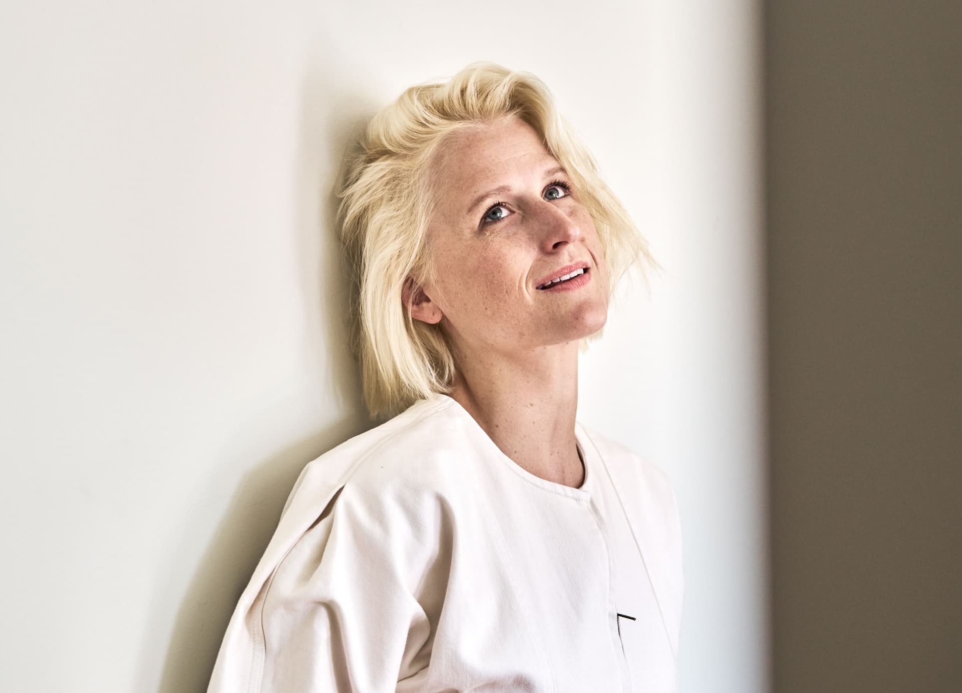 Mamie gummer stands in front of a wall wearing a light top