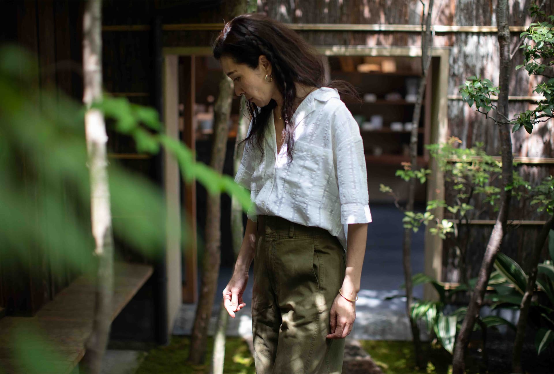 A woman wears a light blouse and green pants while standing outside.