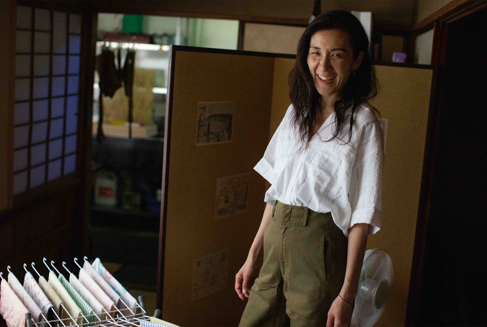 A woman smiles while wearing a light blouse and green pants.