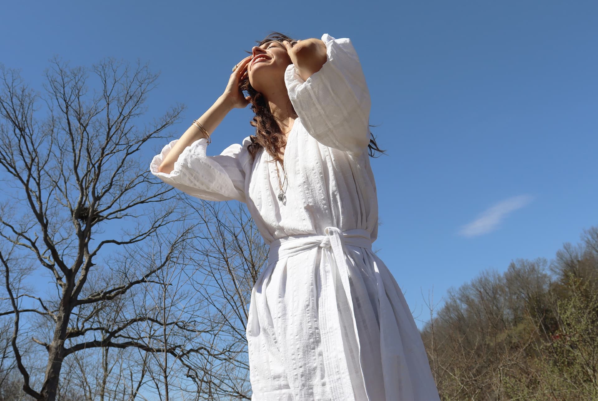 A woman wearing a white dress stands in the sun outside.