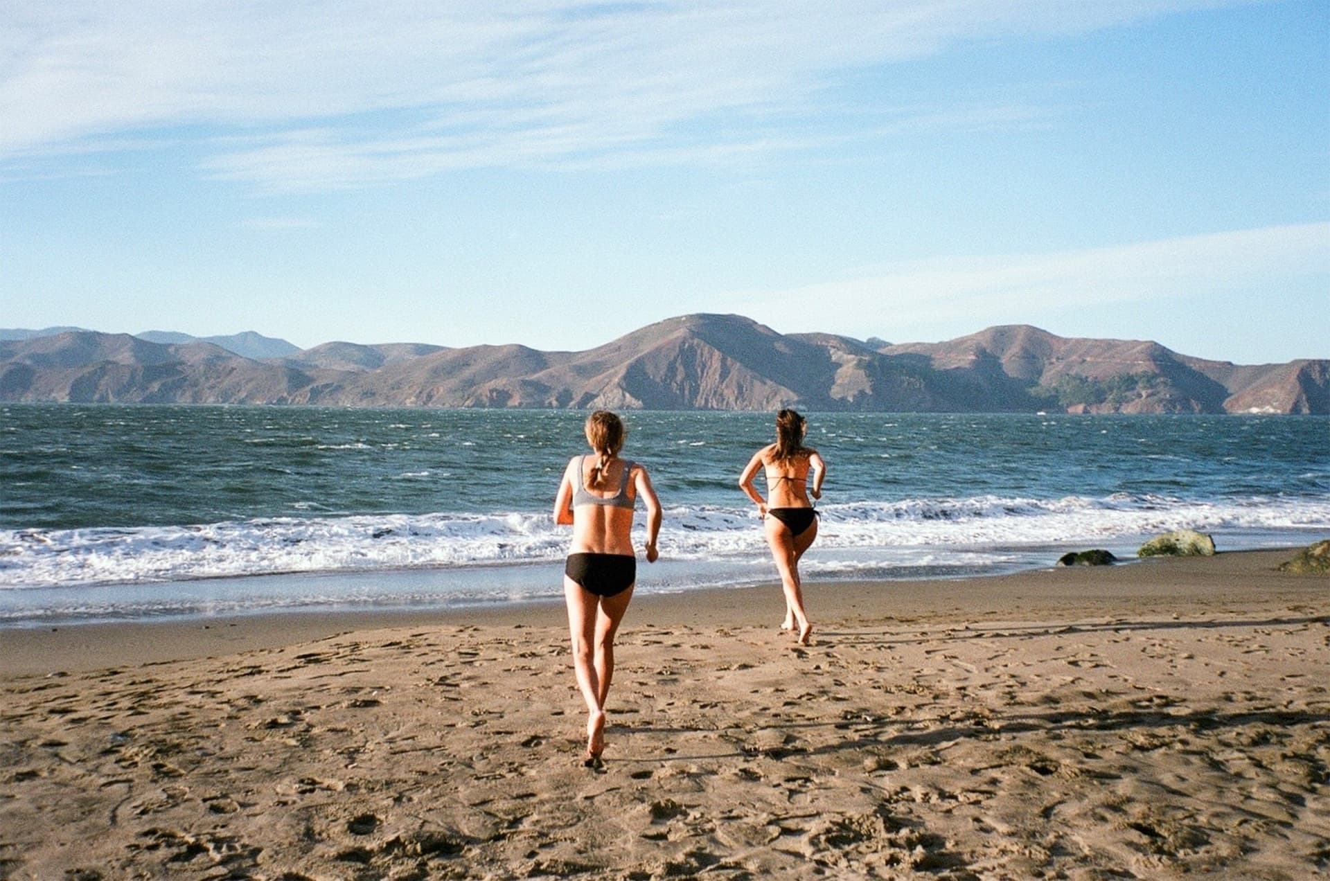 Two women wearing swimsuits run on the beach