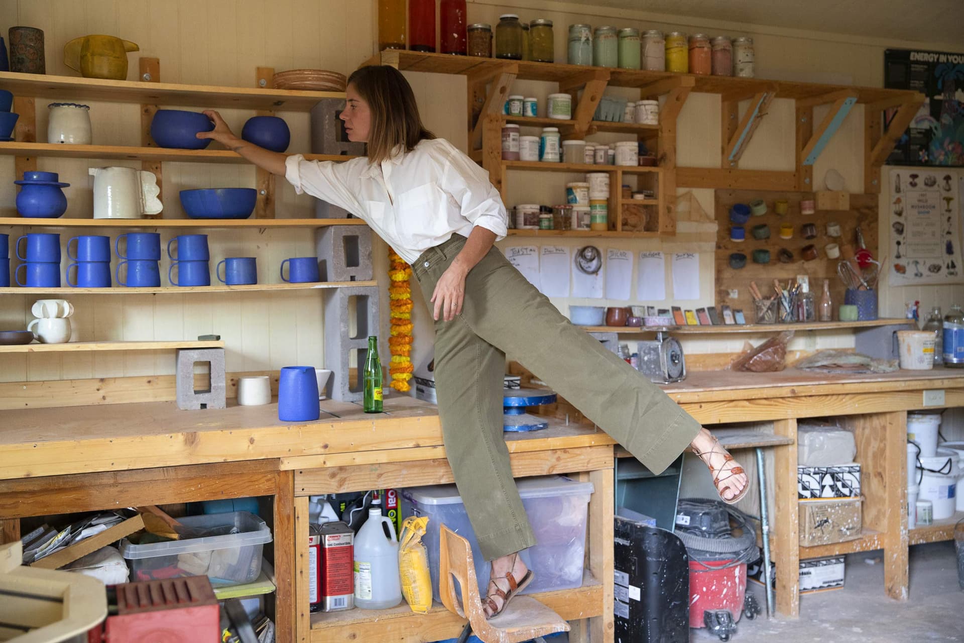 A woman wearing a white shirt and green pants stands on one foot.