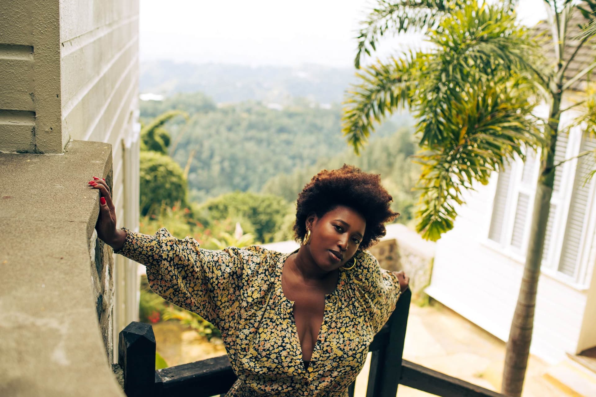 A woman wearing a printed top stands outside and leans against a wall.