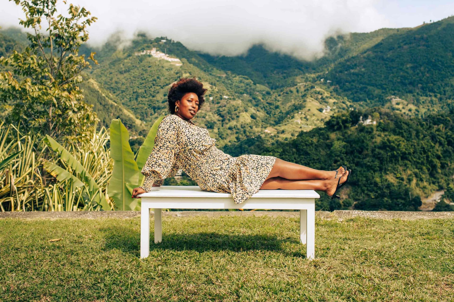 A woman wearing a printed dress lies on a white bench outside