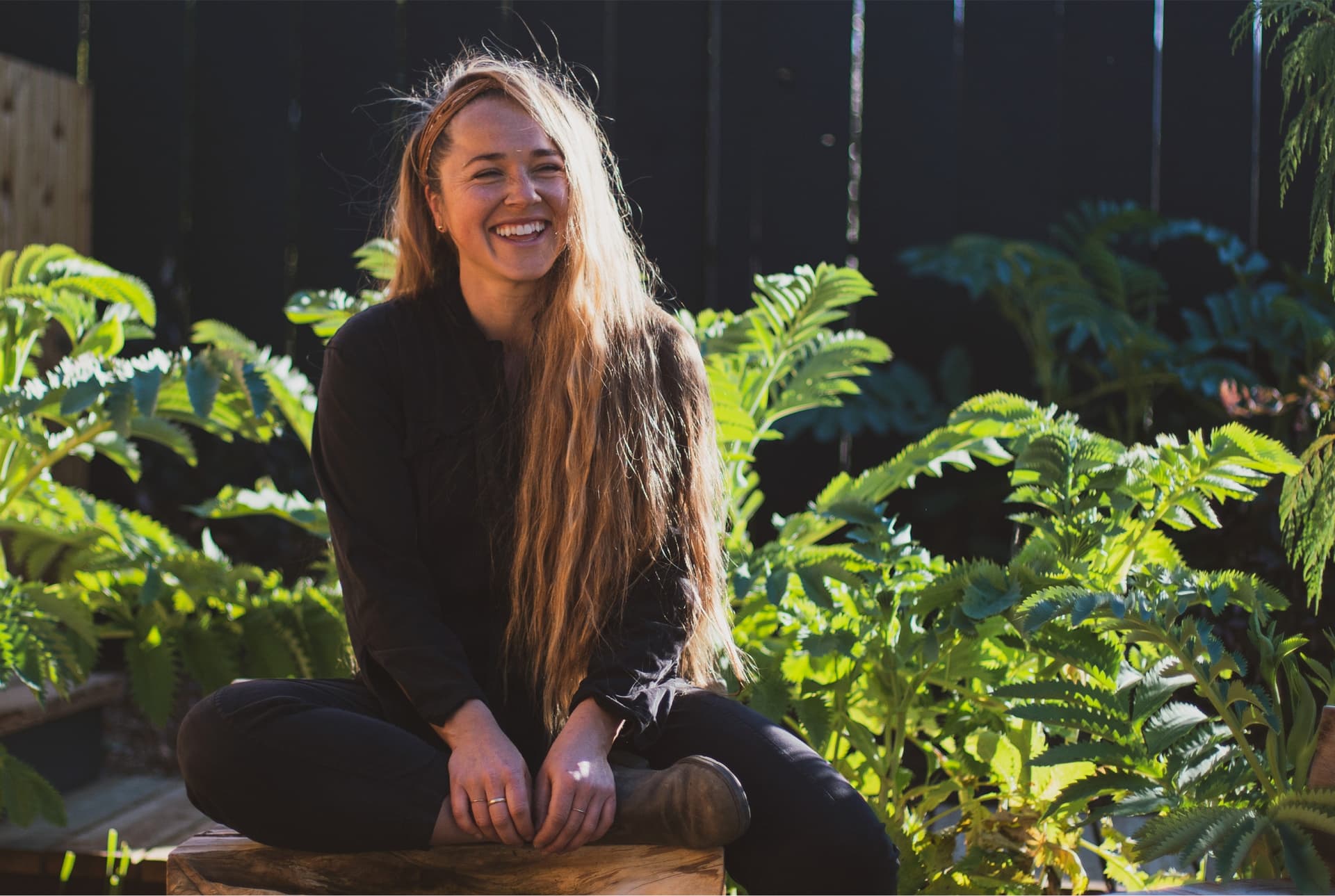 A woman wearing all black sits in front of plants outside.