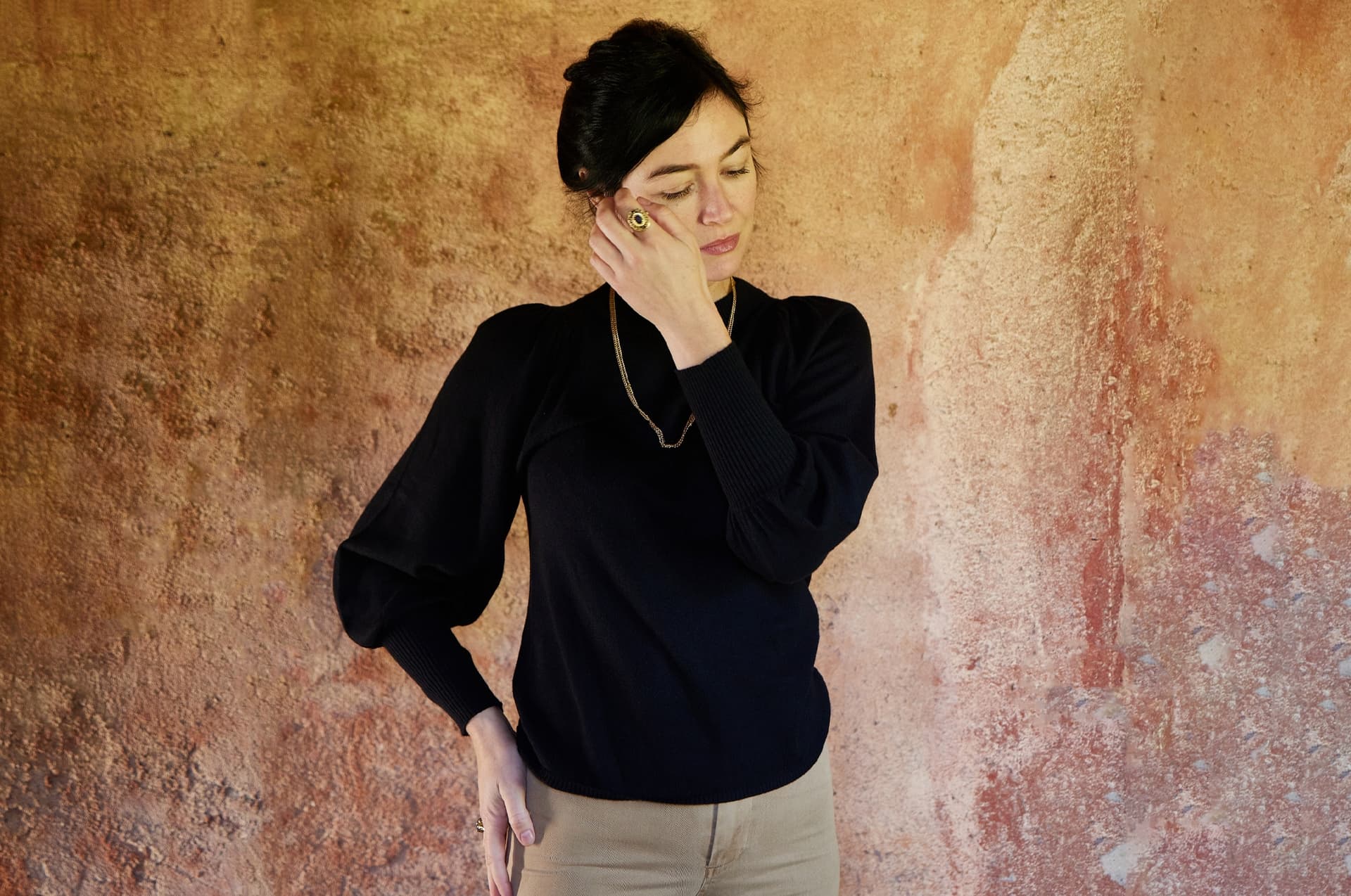 A woman wearing a black sweater stands in front of a brown wall.
